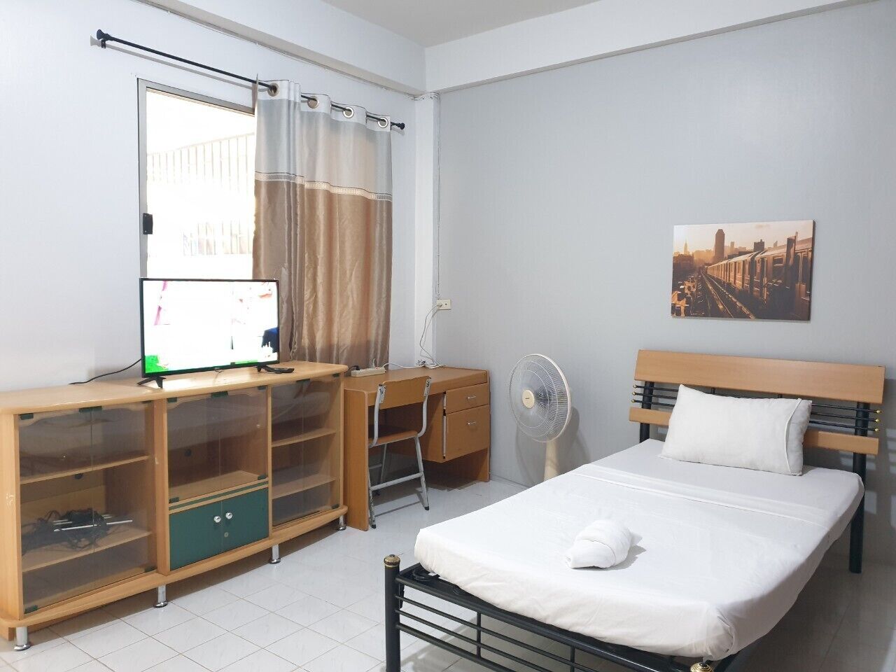 Ein Schlafzimmer mit einem Bett, einem Schreibtisch mit Computer, einem Stuhl, einem Ventilator und einem Fenster mit Vorhängen.