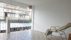 Een ruime kamer met een groot raam dat uitkijkt op een zwembad en een gebouw. Er is een ligbed en een rieten stoel.