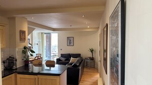 A modern kitchen with wooden cabinets, a black countertop, and a built-in stove. There is a dining area with a black sofa and a small table. The room has a large window and a door leading to an outdoor area.