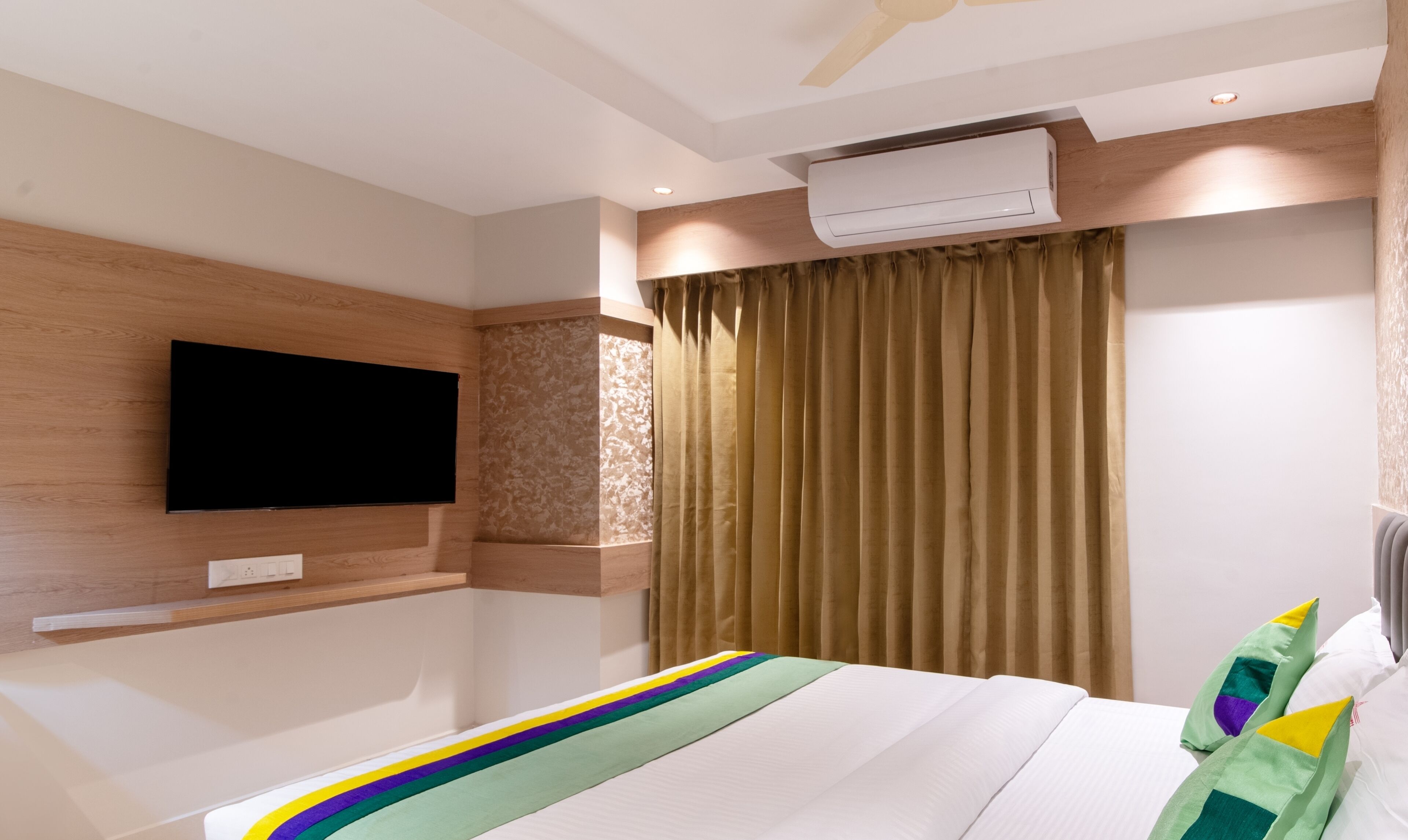 A hotel room with a bed, a mounted TV, an air conditioning unit, and a window with curtains.