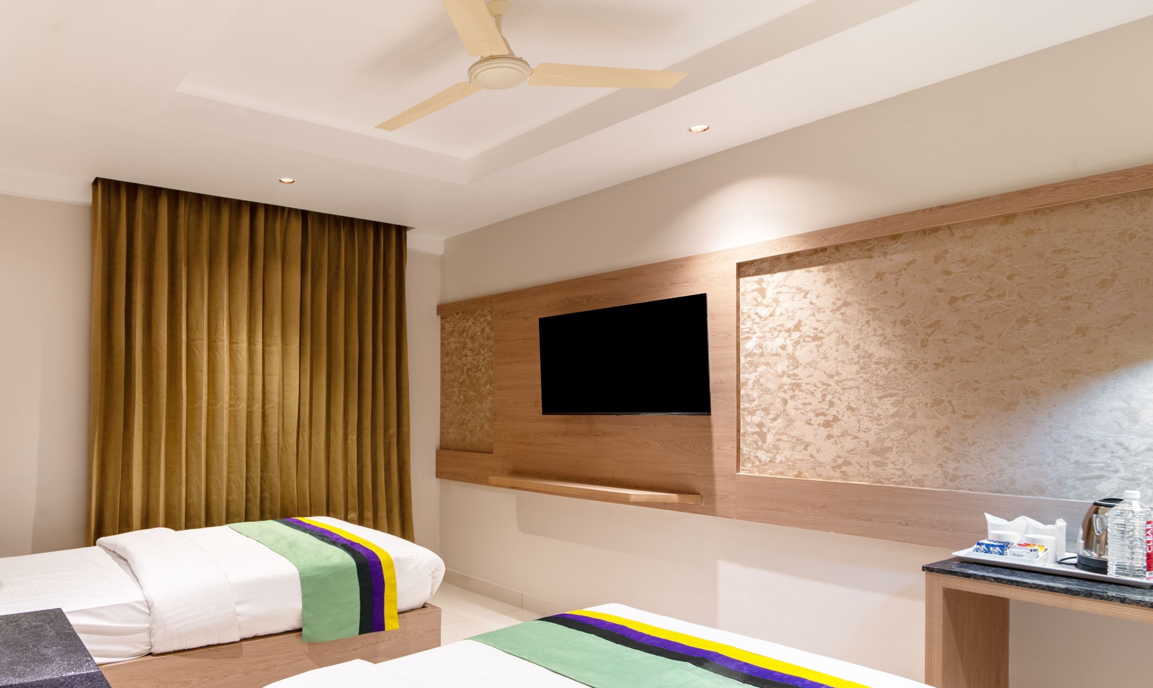 A hotel room with two beds, a flat-screen TV mounted on a textured wall, a ceiling fan, and a small table with toiletries.
