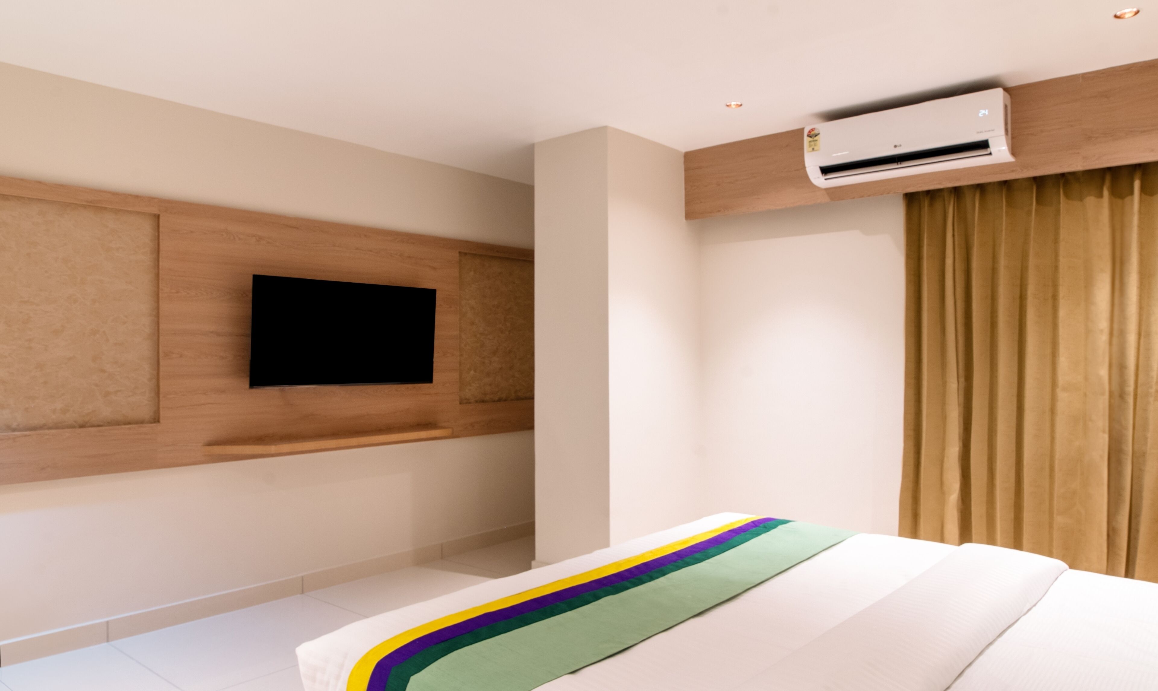 A modern hotel room with a flat-screen TV mounted on the wall, a bed with a striped bedspread, and an air conditioning unit above the bed.