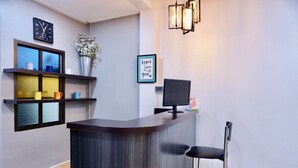 Un espace d’accueil avec un comptoir de réception en bois foncé, un écran d’ordinateur et deux tabourets de bar. On y trouve une horloge au mur, une citation encadrée et un vase avec des fleurs sur une étagère.