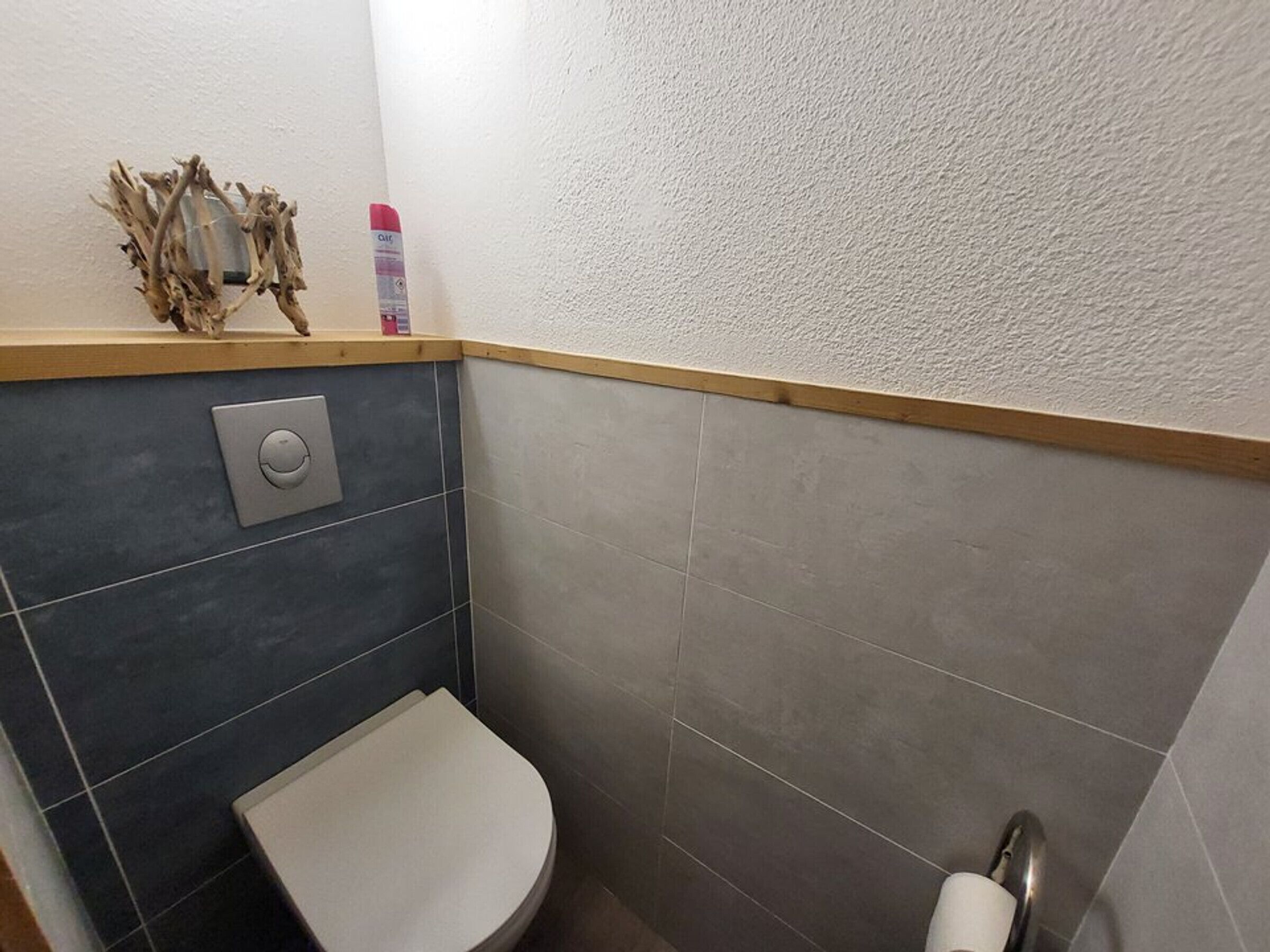Une petite salle de bain comprenant des toilettes, une commande de chasse d’eau fixée au mur et un élément décoratif en bois accroché au mur.