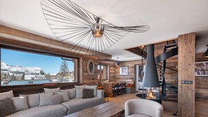A modern living room with a wooden interior, a large window with a view of snow-capped mountains, a fireplace, and a cozy seating area.