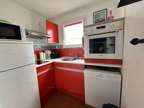 Uma cozinha compacta com micro-ondas, fogão e lava-louças, com armários em vermelho e branco e painel de azulejos.