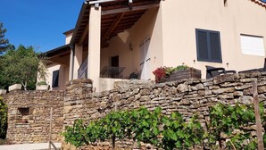 Un muro de piedra con un viñedo al frente, una escalera de piedra que conduce a un porche cubierto, y un edificio con techo de tejas y una pequeña ventana.
