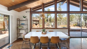 Un comedor espacioso con una gran mesa blanca y cuatro sillas, ventanas amplias que ofrecen vistas al exterior y un techo de madera.