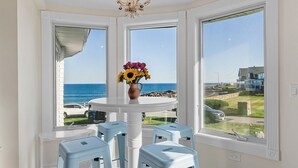 Uma sala de jantar com uma mesa branca e cadeiras azuis, um vaso com flores e vista para o mar através de grandes janelas.