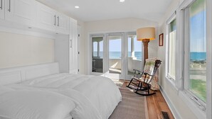 Quarto com cama grande, armários brancos, uma cadeira de balanço e vista para o oceano através de amplas janelas.