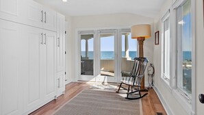 Um quarto amplo com armários brancos, uma cadeira de balanço, um abajur e vista para o oceano por meio de grandes janelas.