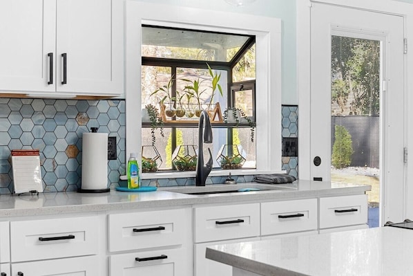 Eine moderne Küche mit weißen Schränken, einem großen Fenster mit Pflanzen und einer Arbeitsplatte mit Kaffeemaschine und Geschirrspülmittel.