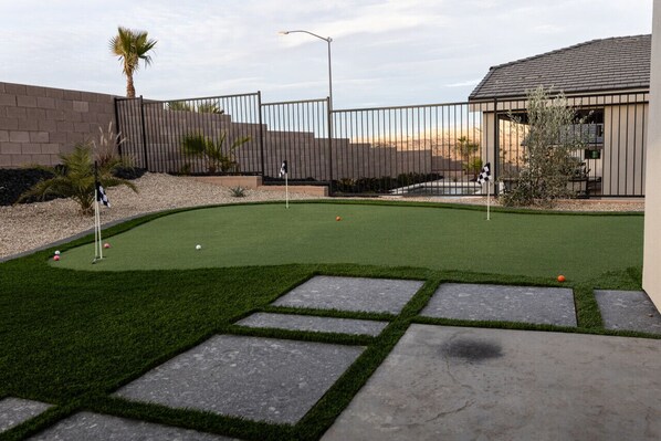 Un campo de golf con césped artificial bien cuidado, senderos de piedra, una cerca y un edificio al fondo.