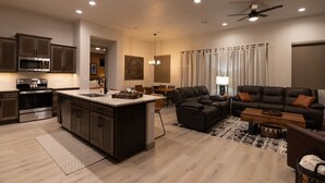 Eine moderne Küche mit integrierten Geräten, ein Essbereich mit einer Pendelleuchte, ein Wohnzimmer mit Sofa und Couchtisch sowie ein Deckenventilator.