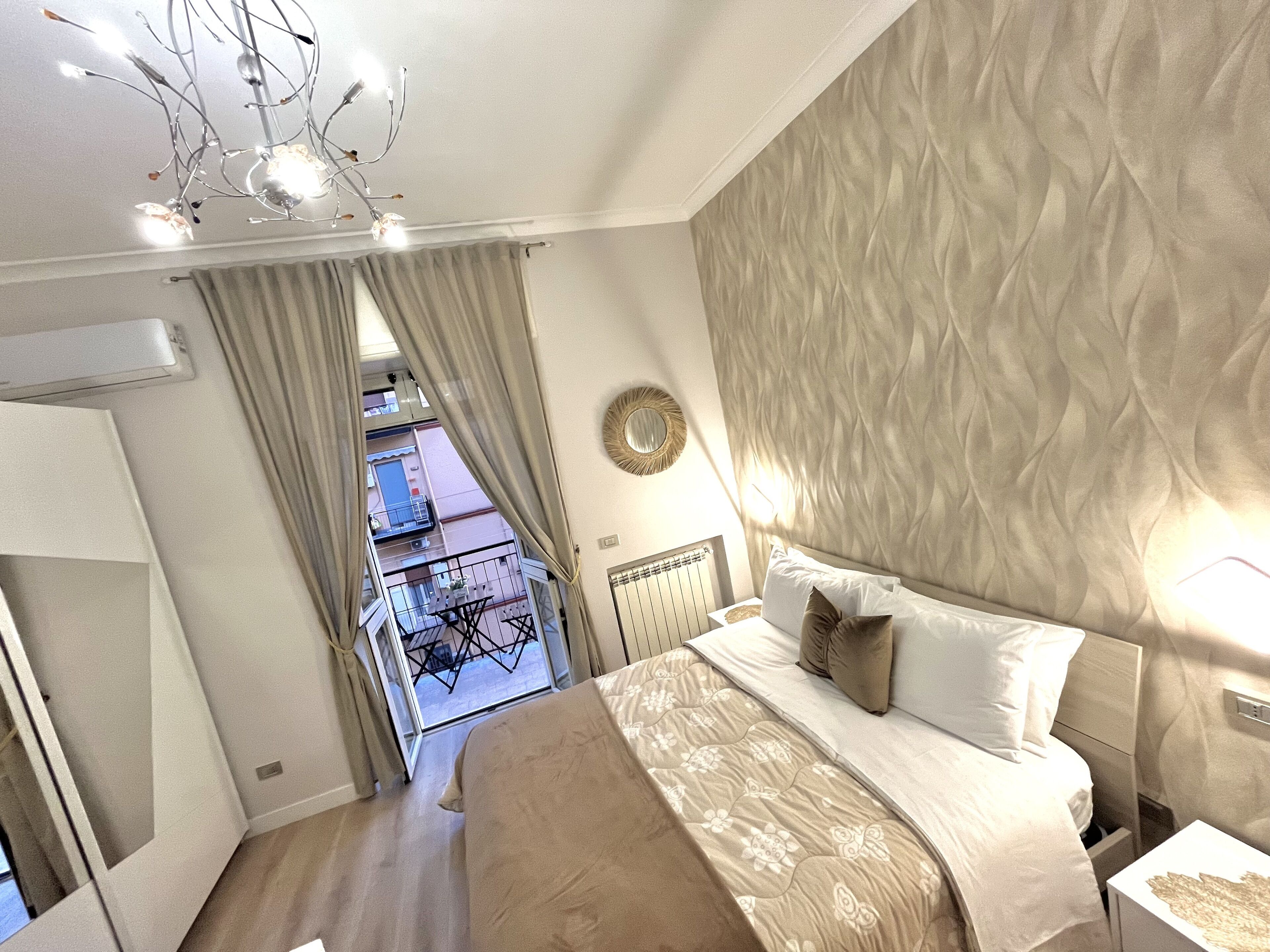 A bedroom with a bed, curtains, a ceiling light fixture, a wall-mounted mirror, and a view of an outdoor balcony.