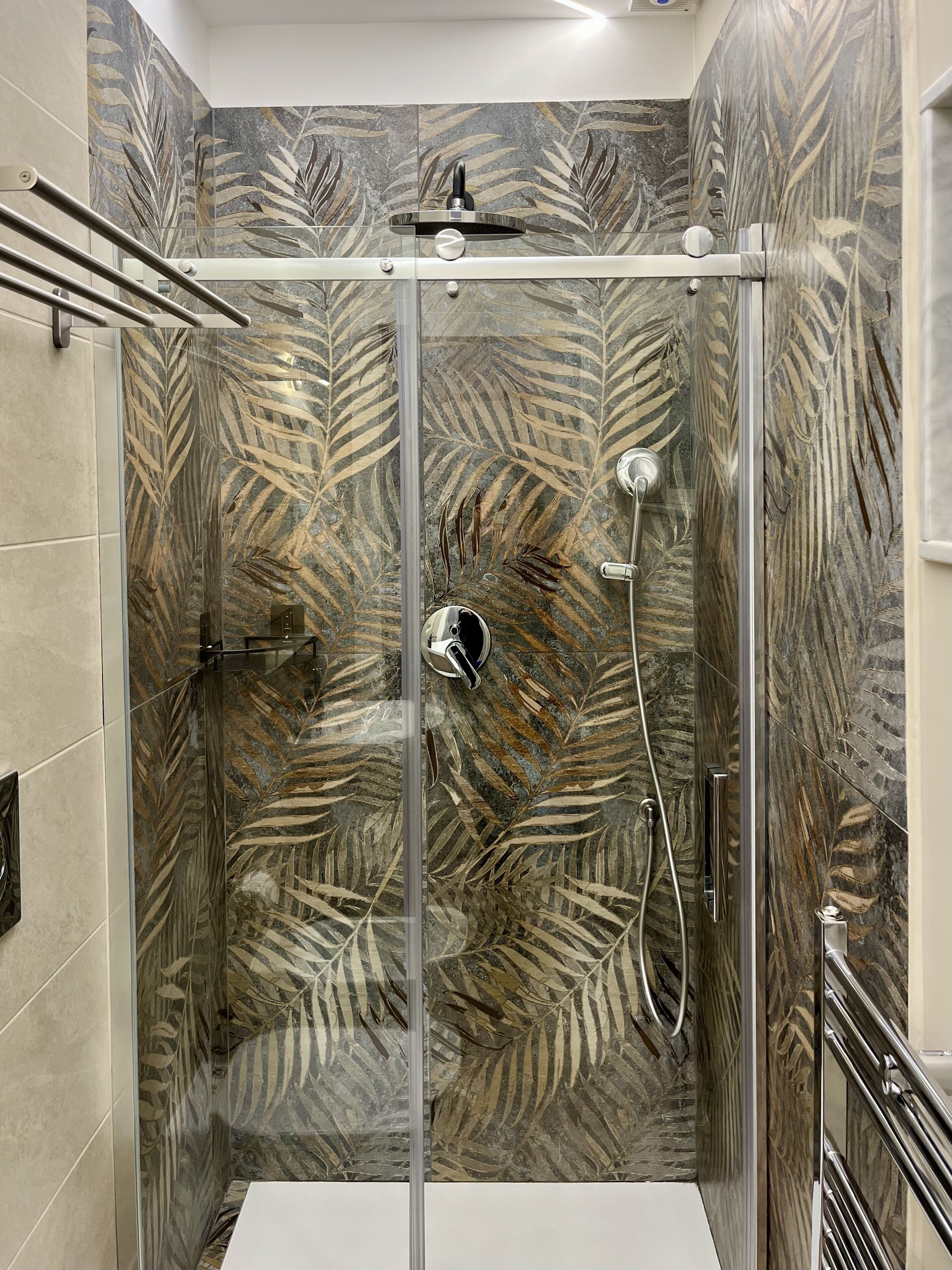 Um chuveiro envidraçado com parede de azulejos com padrão de folhas, um chuveiro manual e uma torneira fixada na parede.