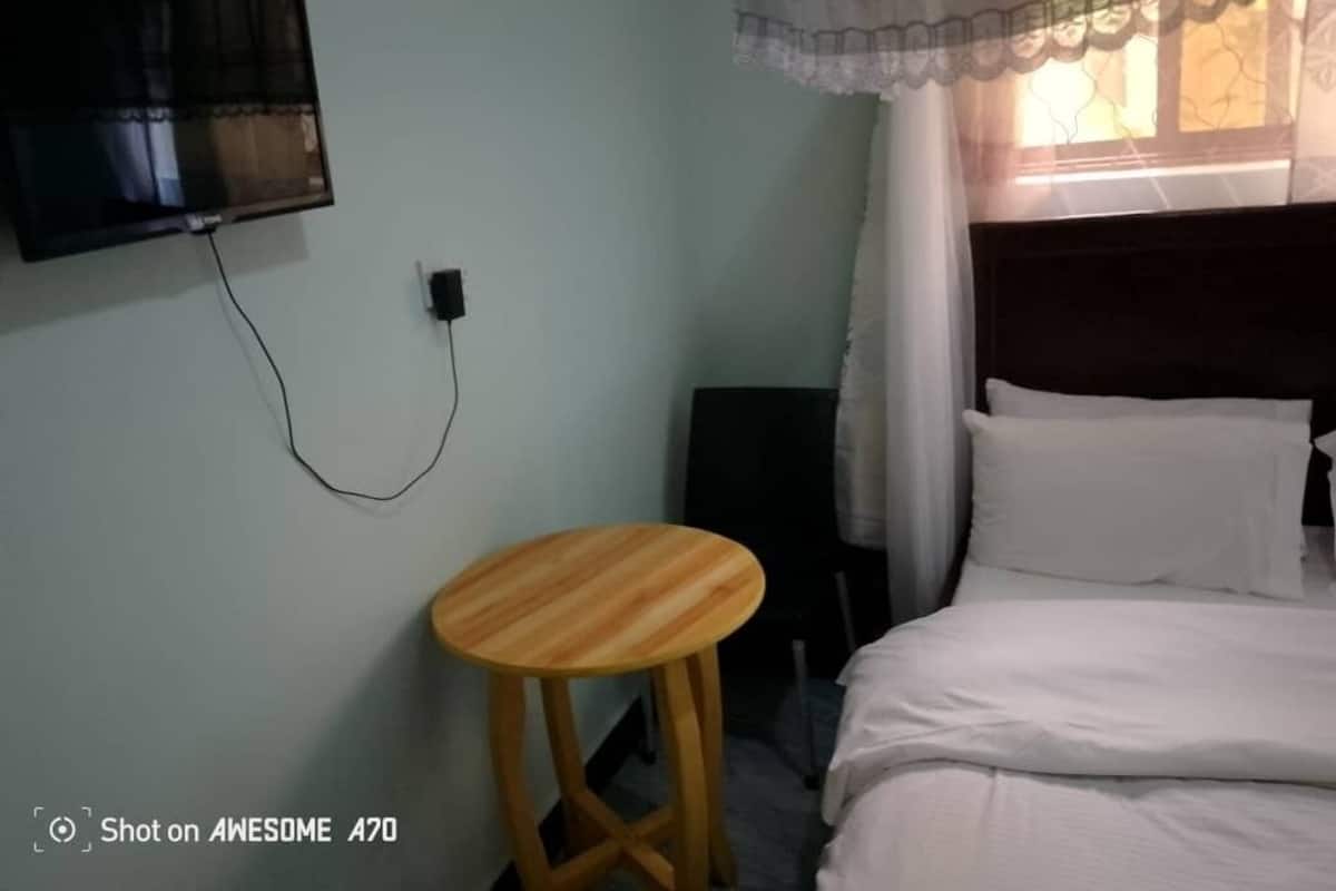 A room with a bed, a television mounted on the wall, a small wooden stool, and a window with a sheer curtain.