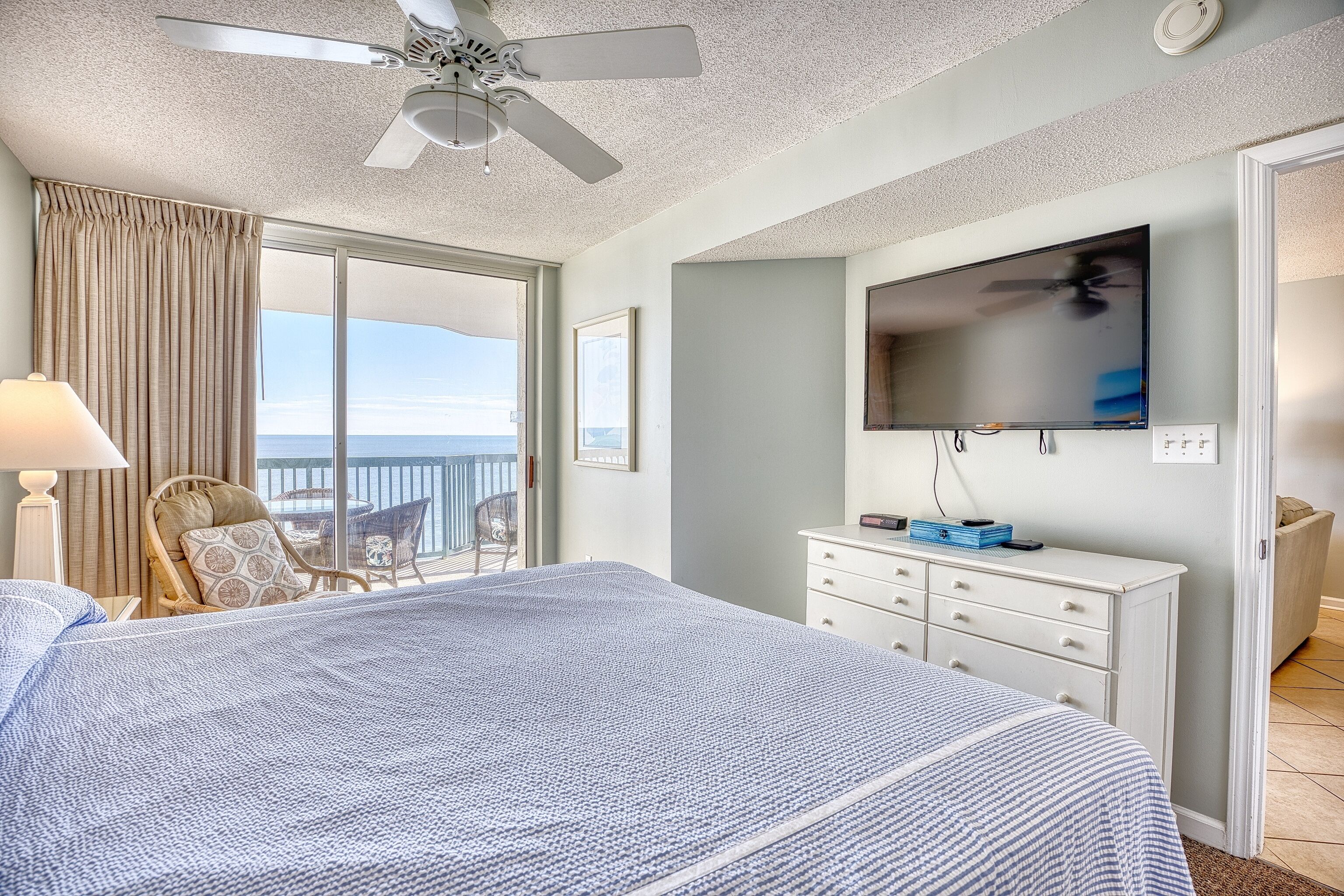 Une chambre à coucher avec un lit, un ventilateur de plafond, une télévision, une commode et une vue sur l’océan.