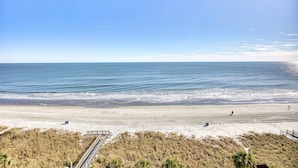 En strand med klart skys og sandet bred, og en træpromenade der fører ned til vandet.