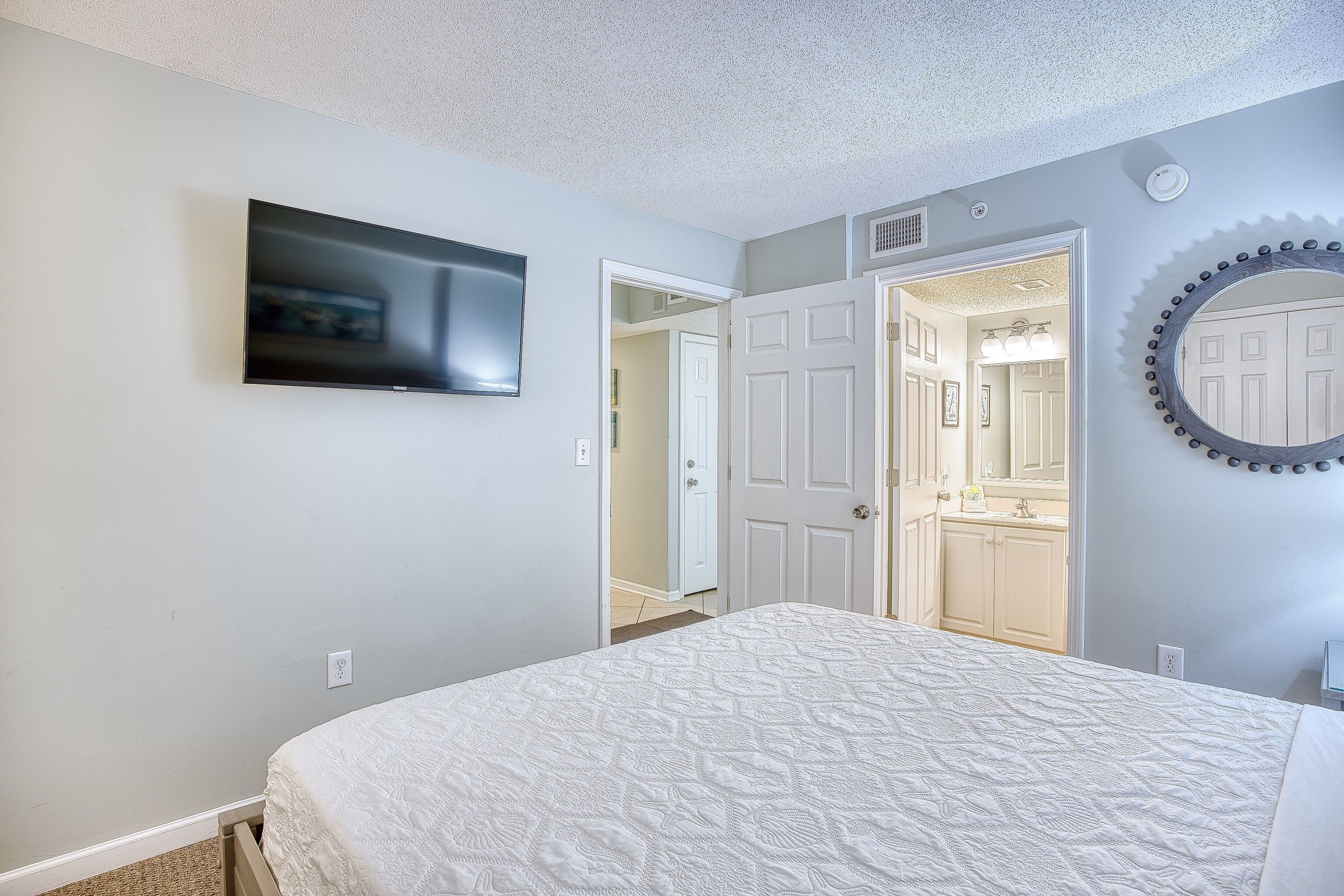 Une chambre à coucher comprenant un lit, un téléviseur fixé au mur, un miroir et une porte donnant sur une salle de bain.