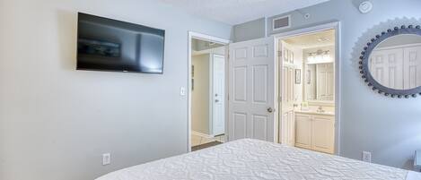 Une chambre à coucher comprenant un lit, un téléviseur fixé au mur, un miroir et une porte donnant sur une salle de bain.