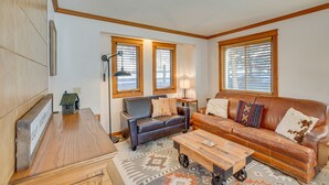 A living room with a brown leather sofa, a wooden coffee table, and a patterned rug. There are two windows with blinds, a lamp, and a small table with a house model. - Spacious Ouray Townhome - Walk to Hot Springs! (Ouray)