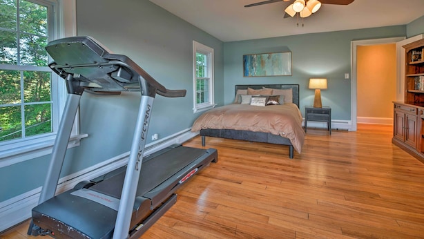 A bedroom with a treadmill, a bed, a ceiling fan, and a window with a view of trees.