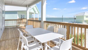A covered outdoor area with a wooden deck, white tables and chairs, and a view of the ocean and beach houses.