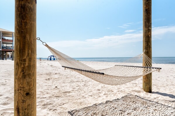 Una hamaca sobre una playa arenosa con postes de madera, cielo despejado y el océano al fondo.