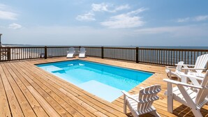 Eine Holzterrasse mit einem Schwimmbecken, weißen Liegestühlen und Blick auf den Ozean.