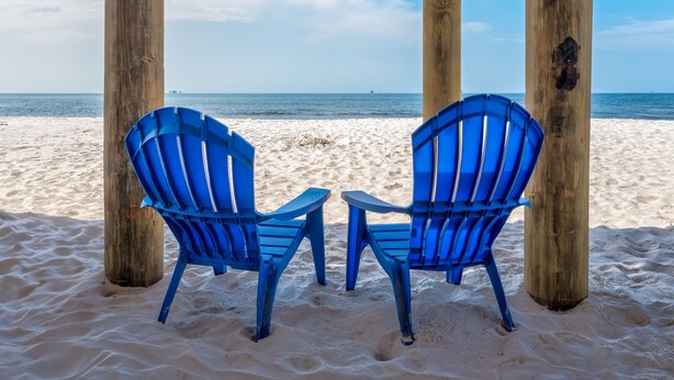Zwei blaue Stühle unter hölzernen Säulen an einem Sandstrand mit Blick auf das Meer.