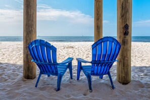 Dos sillas azules bajo pilares de madera en una playa arenosa, con vista al océano.