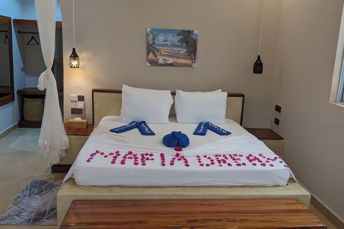Una cama bien hecha con una toalla y otra toalla azul dispuestas en forma de corazón, un cuadro con escena de playa y una mesita de noche con una lámpara.