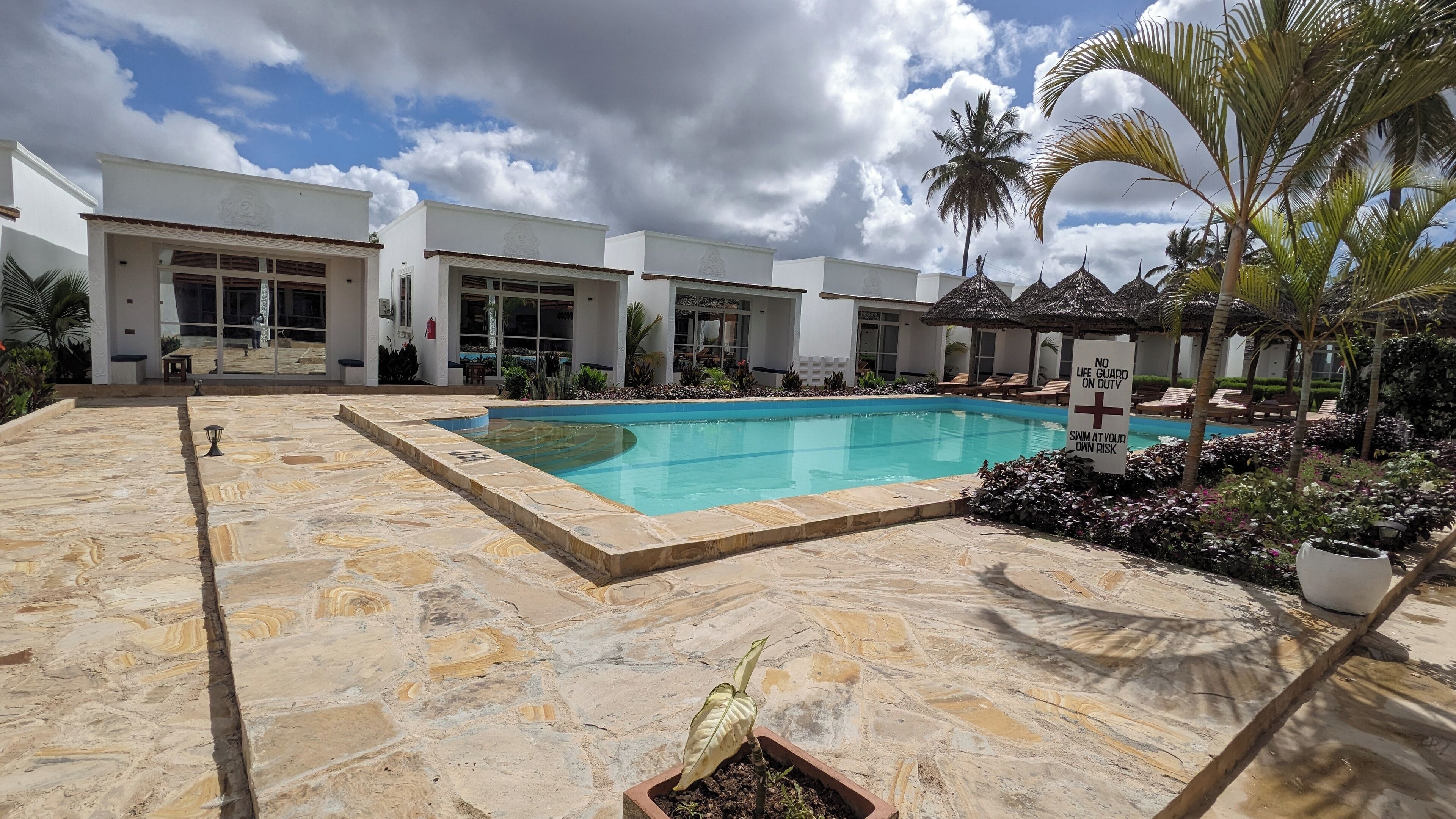Una piscina rodeada por un patio de piedra, palmeras y un edificio con grandes ventanales.