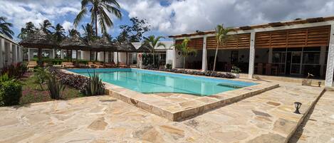 A swimming pool with stone tiles, palm trees, and a covered seating area.