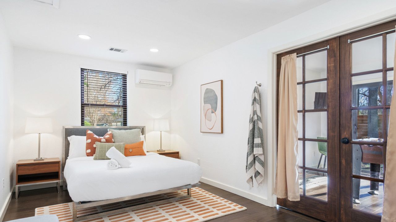 Une chambre à coucher comprenant un lit, des tables de chevet, une fenêtre avec des stores, une porte avec des panneaux de verre et une vue sur l’extérieur.