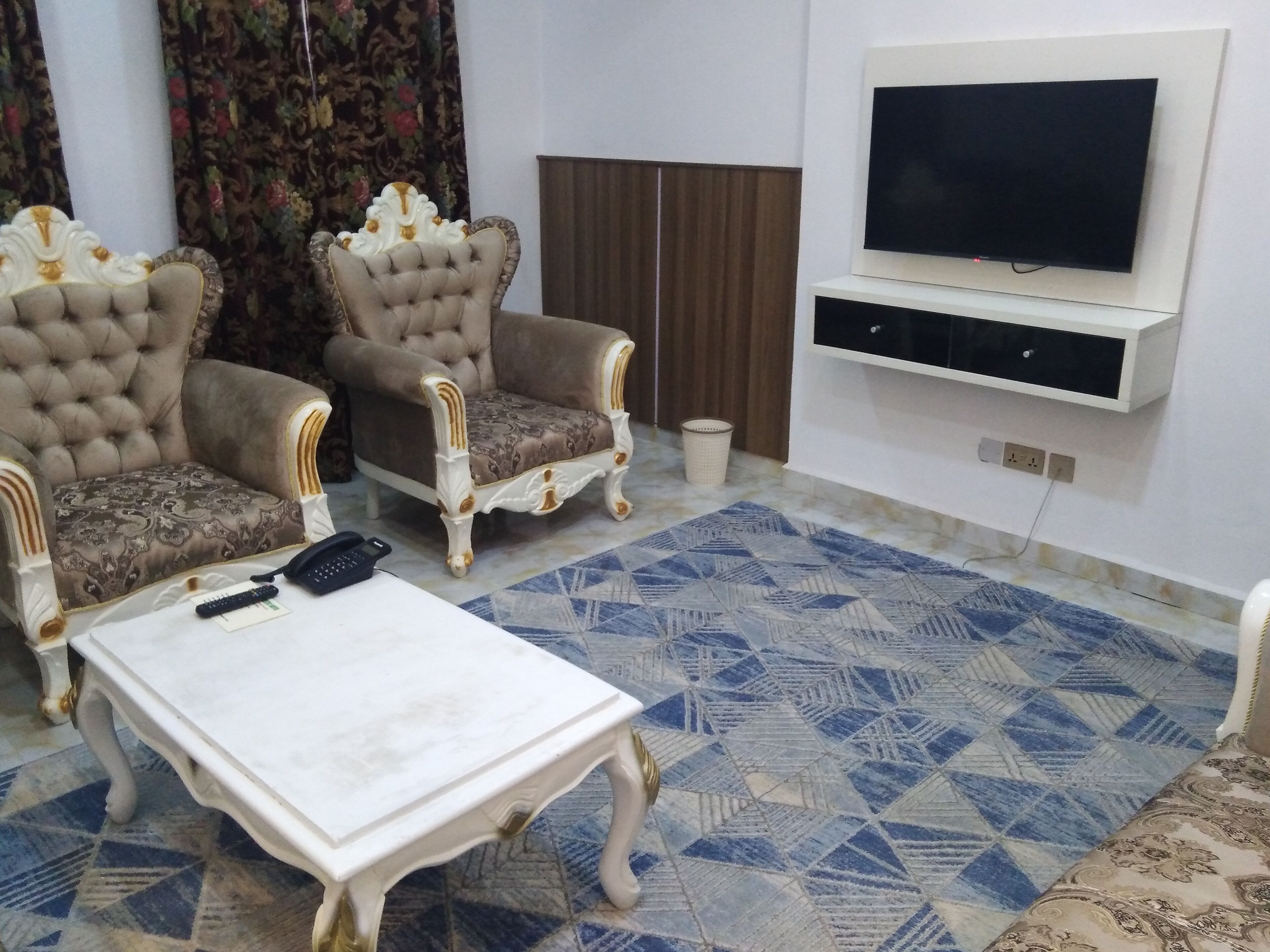 Een woonkamer met twee sierlijke fauteuils, een witte salontafel, een vloerkleed met patroon, een aan de muur bevestigde televisie en een kast eronder.