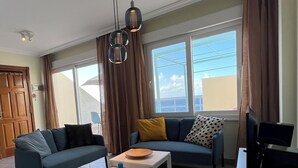 Una sala de estar con un sofá, una mesa de centro y un televisor. Hay cortinas, una ventana con vistas y una lámpara en el techo.