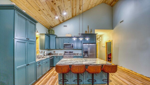 A kitchen with blue cabinets, a marble countertop, a central island with bar stools, and wooden flooring.