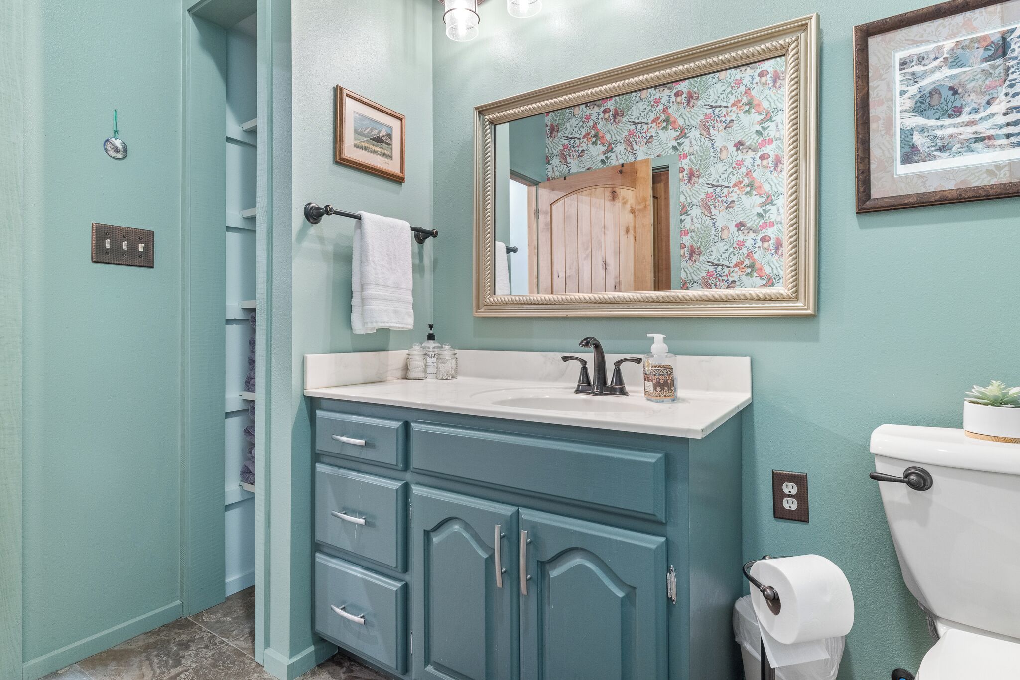 Une salle de bain avec un mur bleu clair, un lavabo blanc avec un meuble-vasque en bois, un miroir avec un cadre floral et des toilettes.