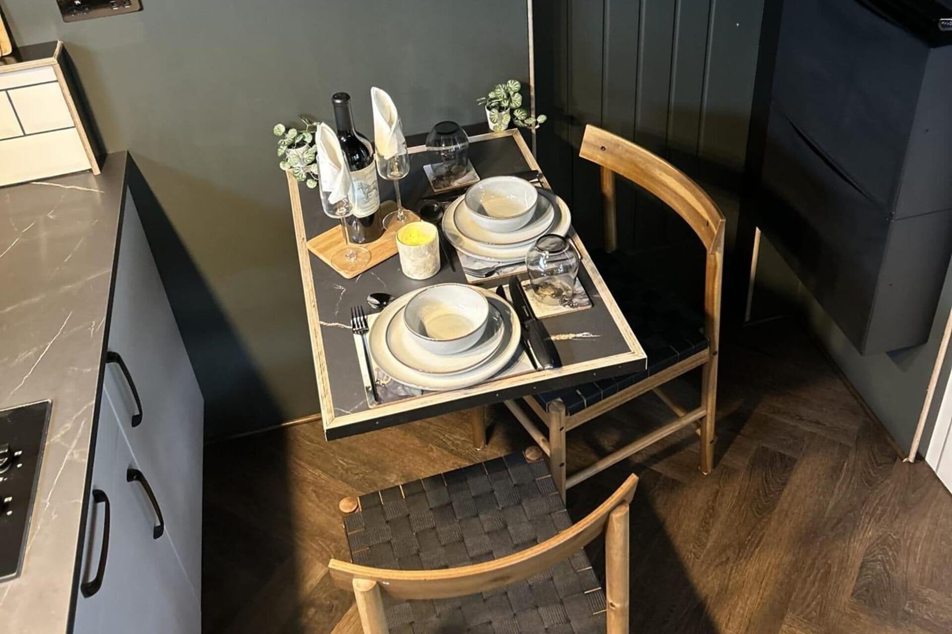 Una mesa de comedor dispuesta con platos, vasos y una botella, en una habitación con piso de madera y una silla.