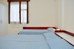 Une chambre avec deux lits, des têtes de lit en bois, des murs blancs et une fenêtre ornée de rideaux légers.