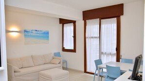 A room with a sofa, a dining table, and chairs. There is a painting on the wall and a window with curtains. - Bright Seafront Flat in Bibione With Side sea View (San Michele al Tagliamento)