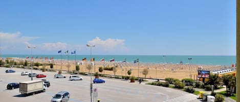 Un estacionamiento con coches y un camión, una playa con banderas y un cielo azul despejado.