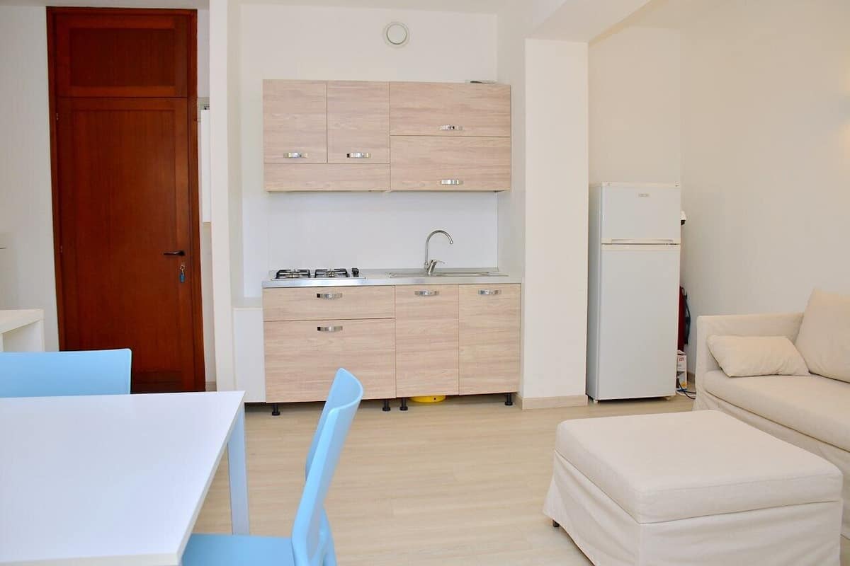 Une cuisine compacte avec des meubles en bois clair, un évier et un réfrigérateur blanc. On y trouve une table à manger avec des chaises bleues et un canapé beige dans l’espace salon.