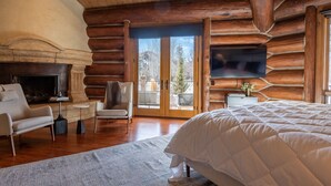 Quarto de cabana de tronco com lareira, uma cama grande, televisão fixada na parede e varanda com vista para árvores e montanhas.