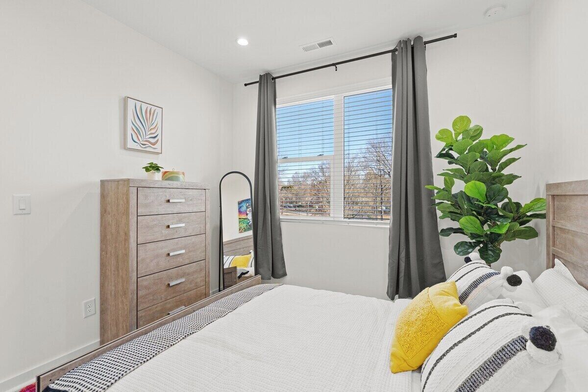 A bedroom with a bed, a dresser, a window with curtains, a plant, and a wall-mounted artwork.