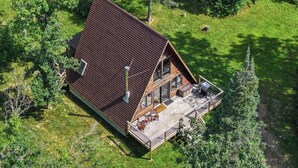 Una cabaña con techo inclinado, una terraza con mobiliario exterior y un ventanal grande con vistas a la vegetación circundante.