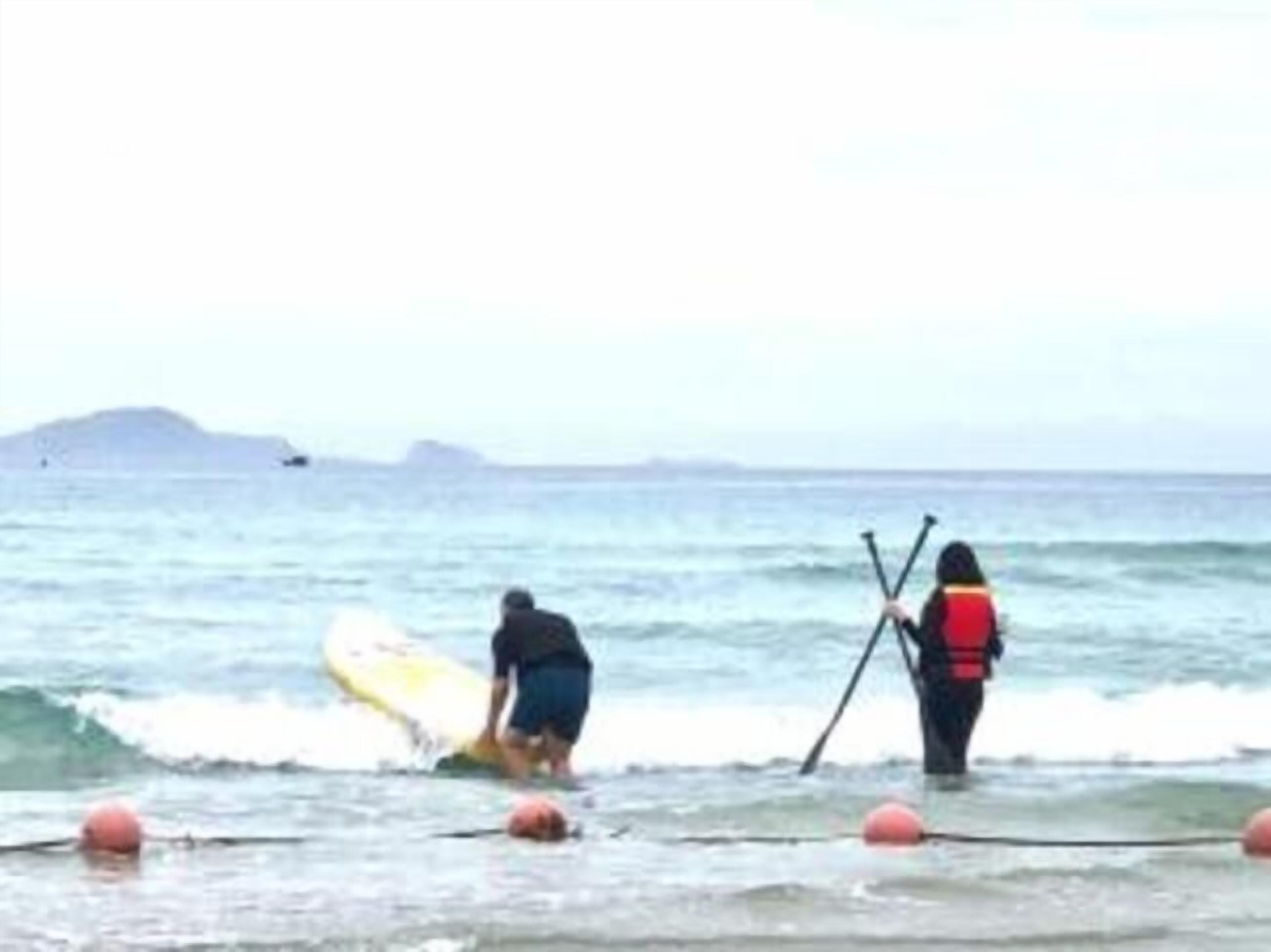 Cena de praia com duas pessoas, uma carregando uma prancha de surf e outra segurando remos, perto da beira da água.