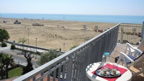 Balcón con una mesa para dos, con vista a una playa que tiene una cerca y cabañas.